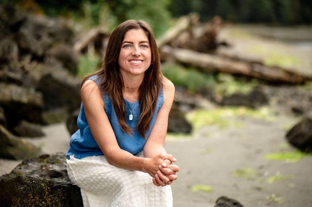 Chrystal smiling while seated on a rocky beach surrounded by coastal greenery.
